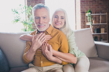 Portrait of attractive elderly careful amorous cheerful couple partners hugging sitting on sofa at home brick loft interior house indoor