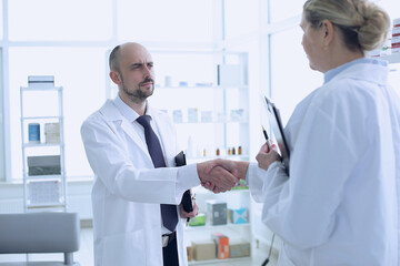 Fototapeta premium Pharmacists is Working. Man and Woman Handshaking with Each Other. Persons is Smiling. People Wearing Special Medical Uniform. People Located in Pharmacy.