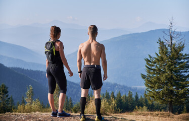 Obraz premium Back view of athletic young man and woman enjoying beautiful view of blue sky and mountains. Sporty couple standing on top of the hill and looking at majestic mountains. Concept of sport, travelling.