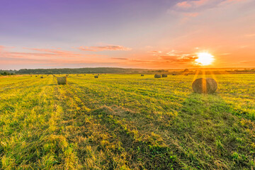 Obraz premium Scenic view at beautiful sunset in a green shiny field in village farm with hay stacks, cloudy sky, golden sun rays, anazing summer valley evening landscape