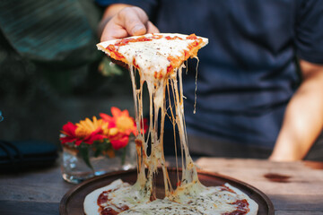 Hand holding homemade mozzarella cheesy pizza on wooden plate at outdoor cafe.
