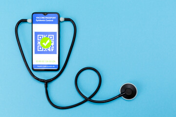 An electronic passport of immunity with a mark on the COVID-19 vaccination on the cellphone screen with a stethoscope and thermometer. Copy space. Blue background. Flat lay