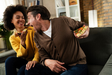 Cheerful young couple sitting on sofa at home. Happy woman and man eating pizza while watching a movie.