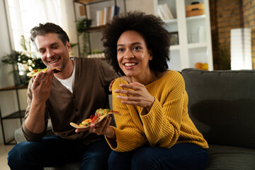 Cheerful young couple sitting on sofa at home. Happy woman and man eating pizza while watching a movie.
