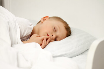 The child sleeps sweetly with his fingers to his mouth on a white crib. A little boy has sweet dreams
