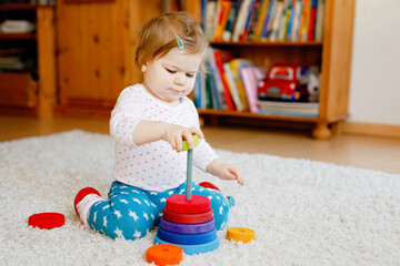 Adorable cute beautiful little baby girl playing with educational wooden toys at home or nursery. Toddler with colorful stack pyramid and music toy. Happy healthy child having fun with different toys