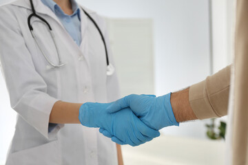 Doctor and patient in protective gloves shaking hands indoors, closeup