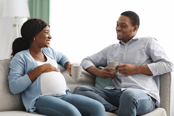 Relaxed expecting black couple sitting on couch, drinking tea