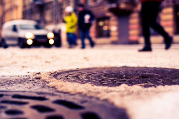 Snowy winter in the big city, pedestrians run across the road in front of riding car. Close up view of a hatches at the level of the asphalt