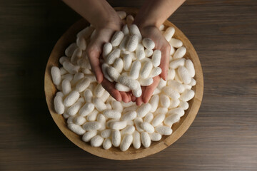 Woman holding white silk cocoons over bowl at wooden table, top view