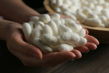 Woman holding white silk cocoons over bowl at wooden table, closeup