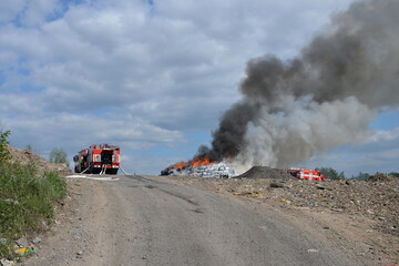 Fire trucks at scene of massive fire in a large landfill with thick black smoke and large flames
