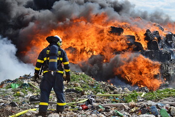 Firefighters extinguish a massive fire in a large landfill with thick black smoke and large flames
