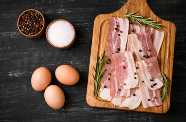 Board with raw bacon with spices, and rosemary on a black background