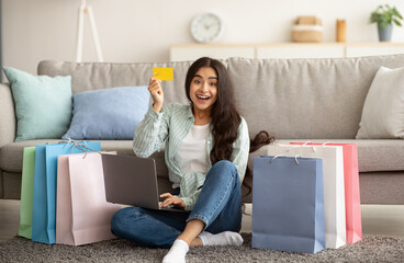 Indian woman feeling happy over sale in online shop, using laptop, showing credit card, sitting on...