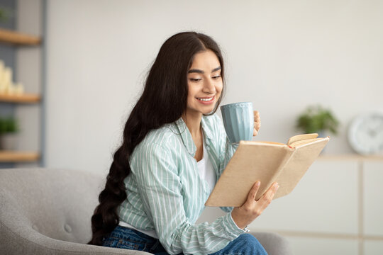 Lovely Indian woman reading book, drinking hot aromatic coffee in armchair at home. Hobbies and pastimes concept