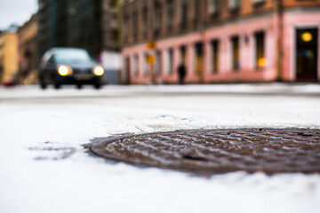 Snowy winter in the big city, the headlights of the approaching car. Close up view of a hatch at the level of the asphalt