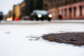 Snowy winter in the big city, the headlights of the approaching car. Close up view of a hatch at the level of the asphalt