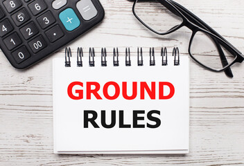On a light wooden table a calculator, glasses and a blank notepad with the text GROUND RULES....