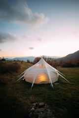 Night time sunset shot of glamping camping tent set up in beautiful nature landscape. Cosy and warm big tent lit from inside with orange dimmed lights. Romantic weekend getaway for couple