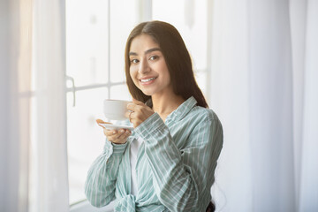 Lovely young Indian woman having coffee break in front of window at home