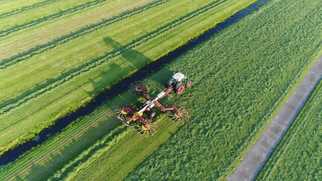 Aerial footage of tractor raking grass flying back right of agricultural machine forming windrow rows of hay raked up to dry before being baled or stored also showing long shadows from sundown 4k