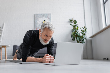 bearded man in wireless earphones doing plank on fitness mat near laptop in living room.
