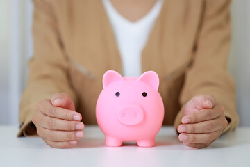 Young active business woman hands in casual dress sitting and protecting piggy bank on white table. Insurance and security concept.