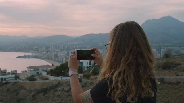 Woman makes photos and videos on her smartphone of cinematic sunset over beautiful city on seaside. Social media content creator or travel blogger