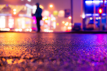 Rainy night in the big city, illumination lights of the shopping center with a man standing on the road. Close up view from the asphalt level