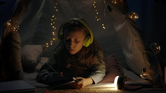 Pretty girl in headphones thinking and writing while lying on floor in decorative makeshift tent at home. Teen listening to music while spending free time in evening. Concept of leisure.