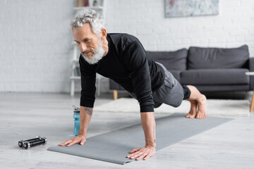 Fototapeta premium tattooed man with grey hair doing plank on fitness mat near dumbbells and sports bottle in living room.