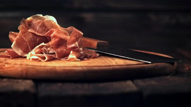Super slow motion sliced into thin pieces of ham falls on a cutting board with a knife.Filmed at 1000 fps. On a wooden background. 