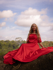 Bali trend photo. Caucasian woman in long red dress sitting on big stone in tropical rainforest. Vacation in Asia. Travel lifestyle. Breathtaking view. Bongkasa, Bali, Indonesia