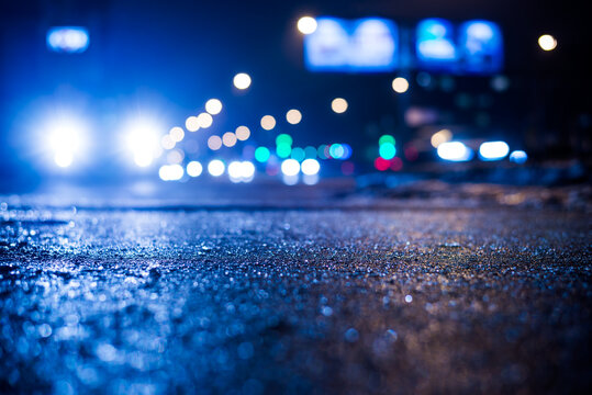 Rainy night in the big city, glare from the headlights of the parked car and a passing near the stream of cars. Close up view from the sidewalk level