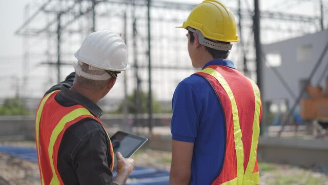 back view of senior contractor manager talking to young asian builder with digital tablet at construction site outdoors . group of diversity construction worker in hard hat meeting outside