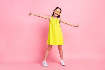 Full length photo of adorable funky school girl wear yellow dress eyewear smiling dancing arms sides isolated pink color background