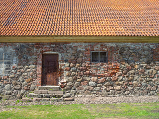 An old wall of a medieval building, patched up with bricks with a rusty iron door, a small window and a new roof. Old building in sunny weather.