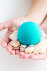 Close-up of blue bubbling round bright bath bomb in the shape of a ball and decorative seashells in woman's hands isolated on a white background. Turquoise aroma bath salts for spa and relaxation