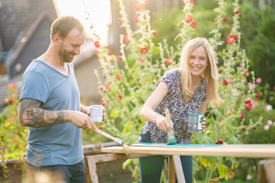 Smiling blond hair woman painting wooden plank with man standing in garden