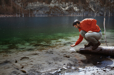 Male tourist crouching on log with water in cupped hand at Lake Kaindy