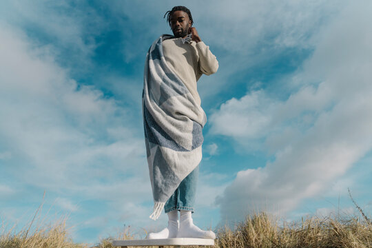 Young man wrapped in blanket standing on stool against cloudy sky
