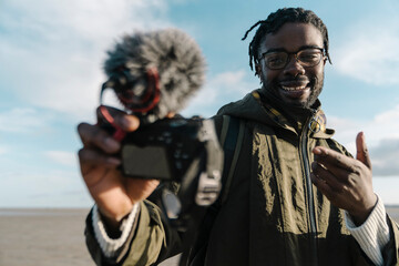 Smile male influencer gesturing while vlogging through camera at beach against sky