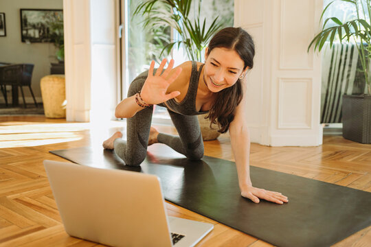 Smiling beautiful female instructor waving during online yoga class through laptop