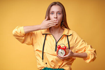 Too late! Young shocked woman  holding an alarm clock on a yellow background.