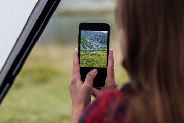 Girl taking photos of the rocky mountains and lake using the phone. Close-up shot of the handі holding the phone. View from the tent.