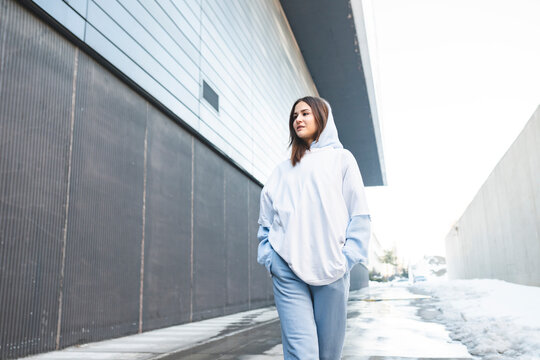 Young woman wearing hood clothing walking with hands in pockets on footpath