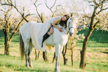 Bride in white dress sits horseback on horse and embraces it in spring blooming garden.
