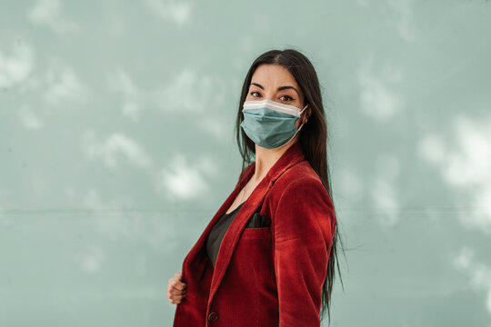 Businesswoman in red blazer wearing protective face mask against turquoise wall during pandemic