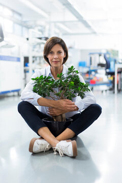 Mature female entrepreneur embracing potted plant sitting in laboratory